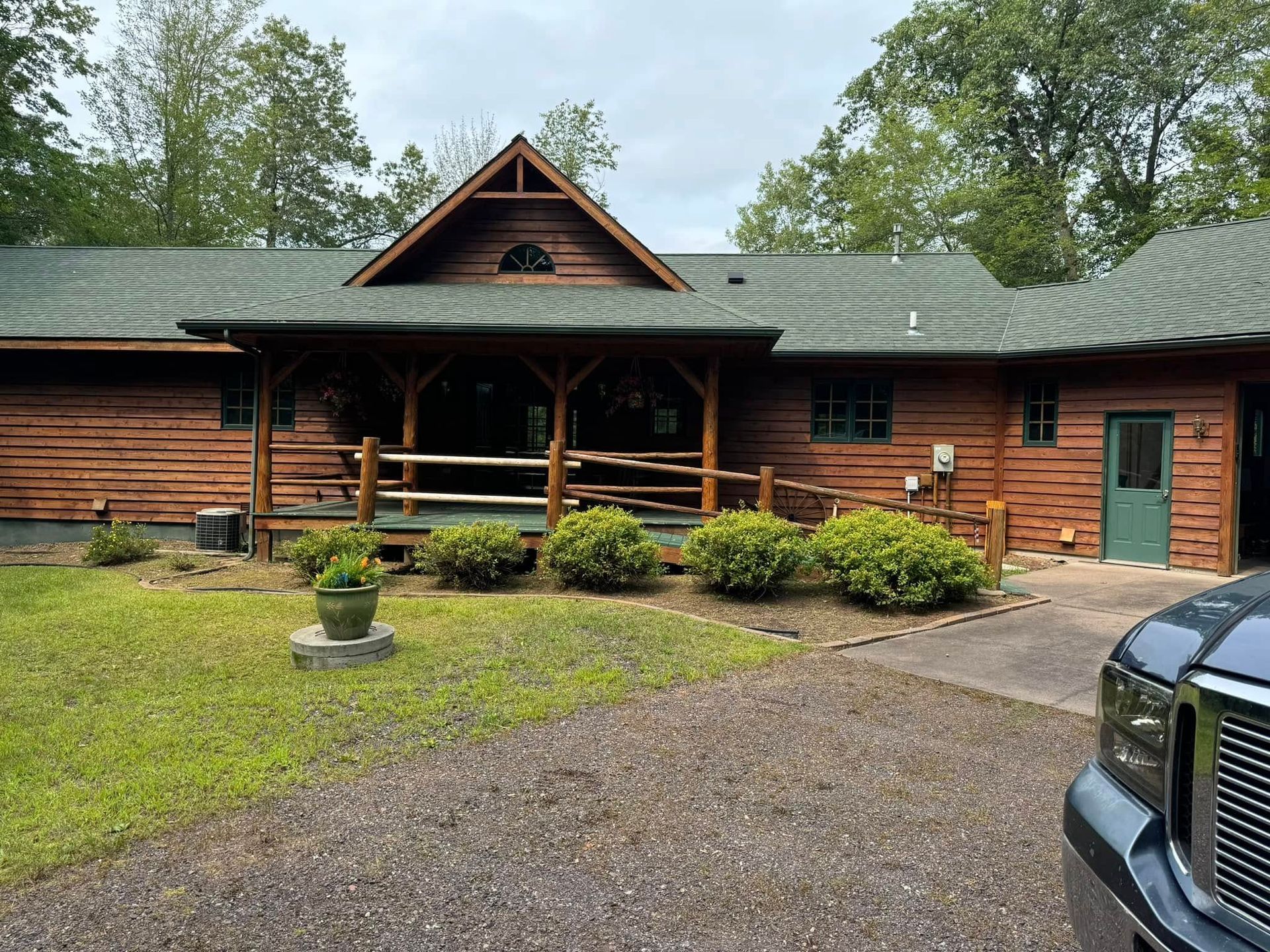 Rustic log cabin with a green roof, porch, gravel driveway, and surrounding trees