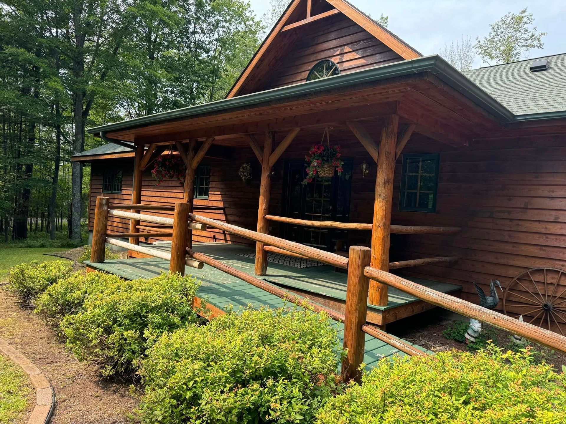 Wooden cabin porch with log railings, green shrubs, and a forested path in front