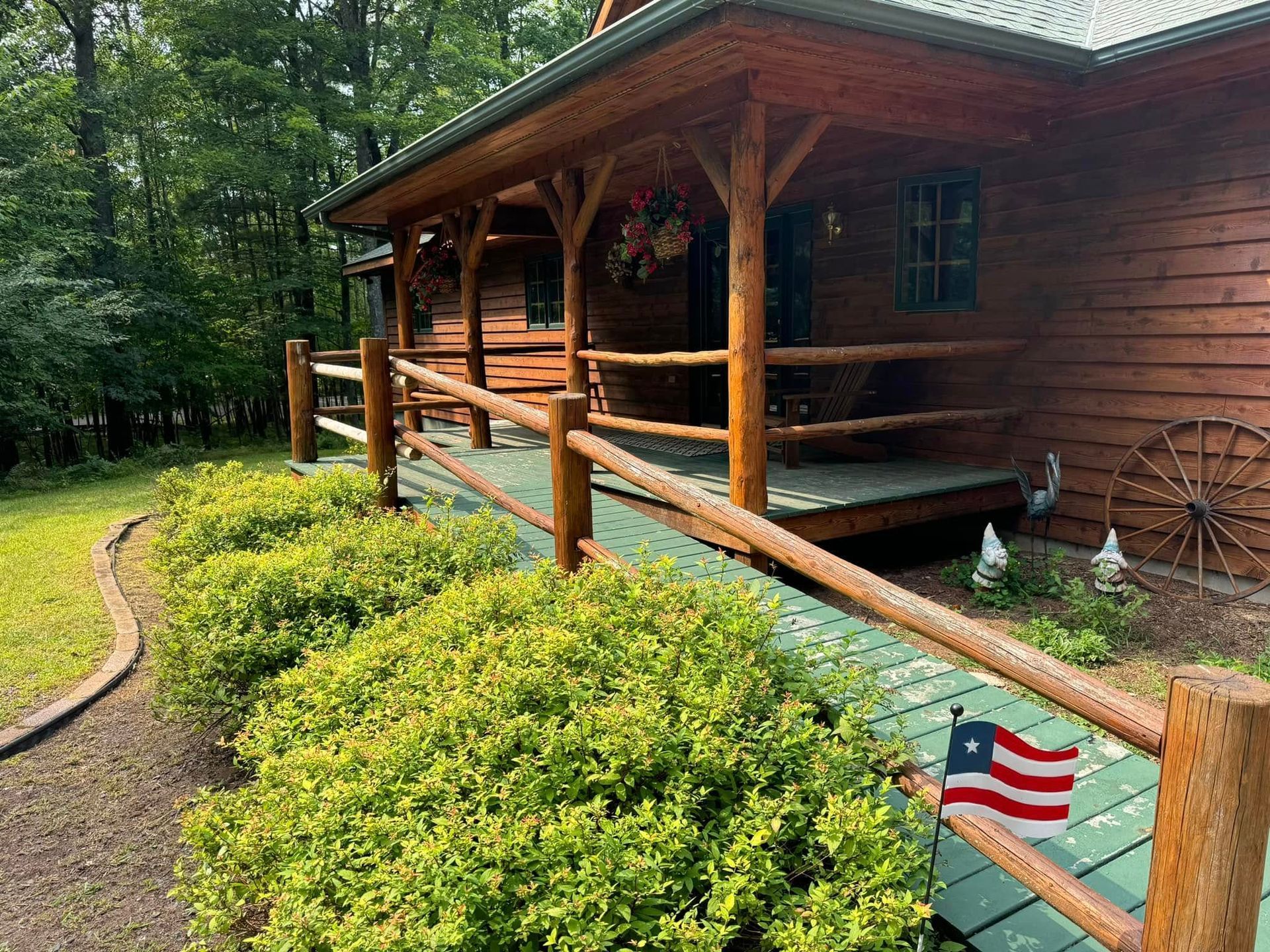 Wooden cabin porch beside a wooded path, with American flags and green shrubs in the foreground