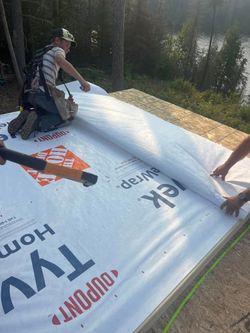 Two workers installing a white roof underlayment on a sloped roof in bright sunlight.
