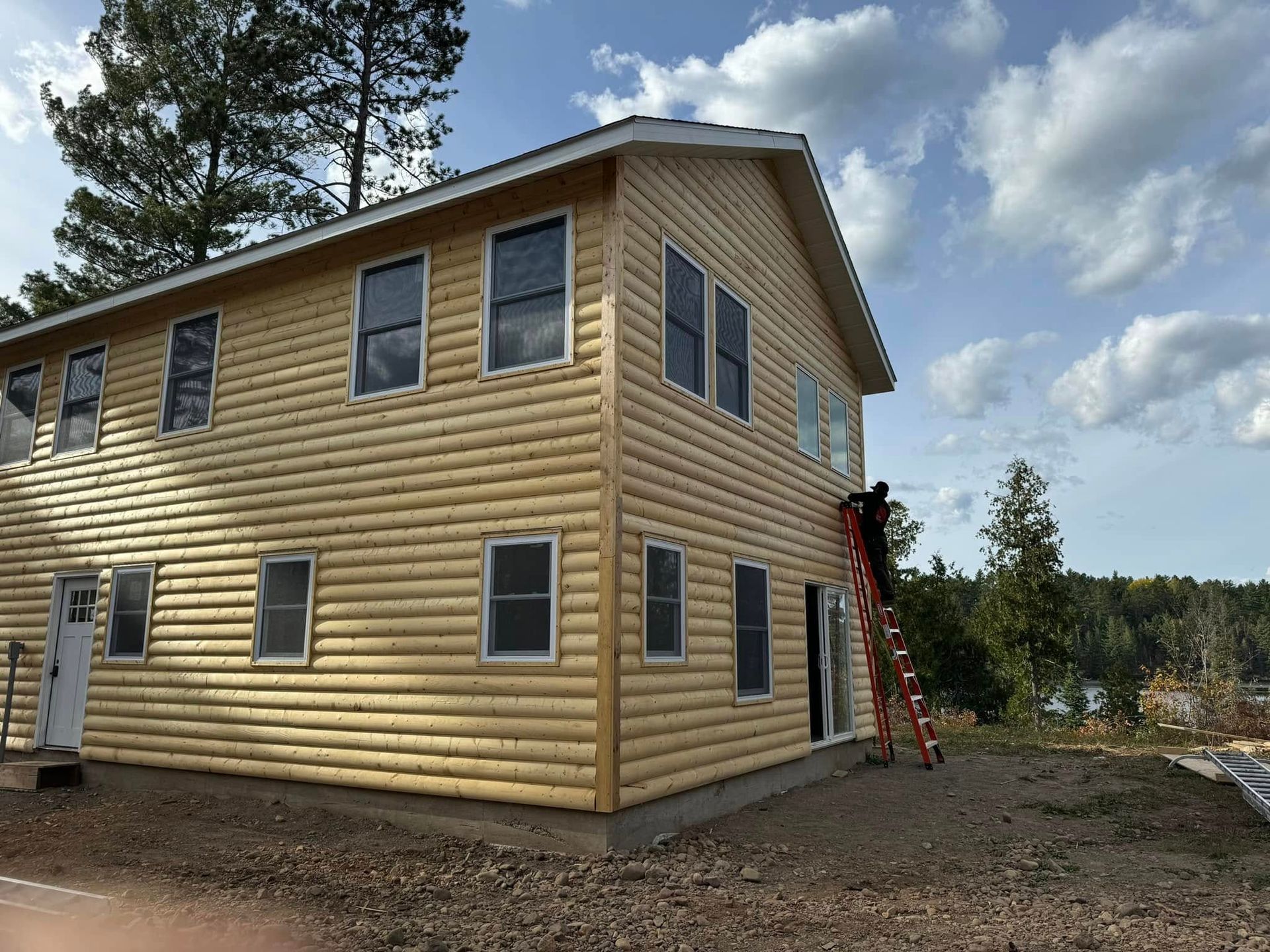 Two-story yellow house with blue trim and a red ladder on the side, set on a wooded lot under cloudy skies