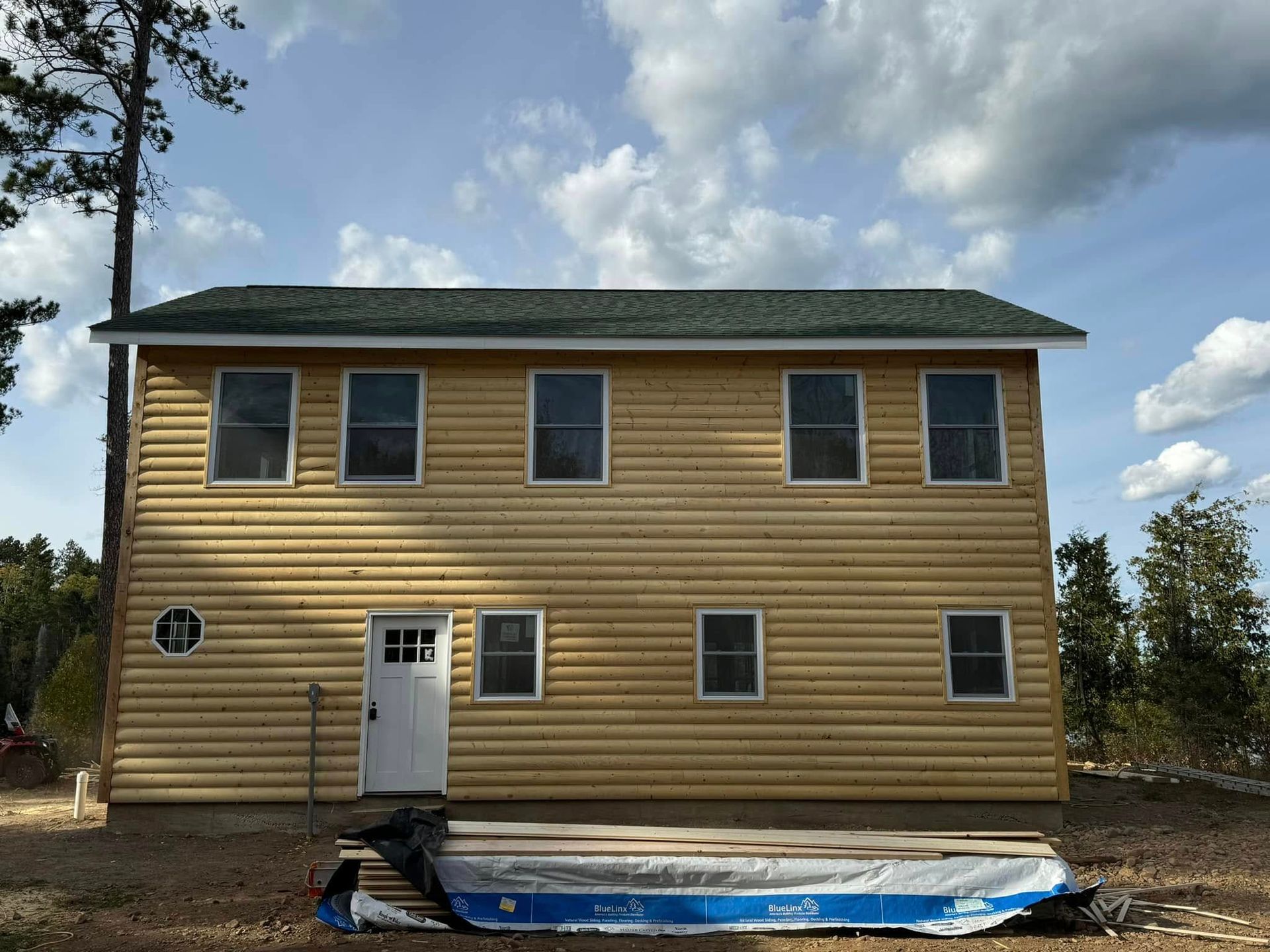 Two-story tan house with green roof under cloudy sky, front door and windows, blue tarp on ground in front