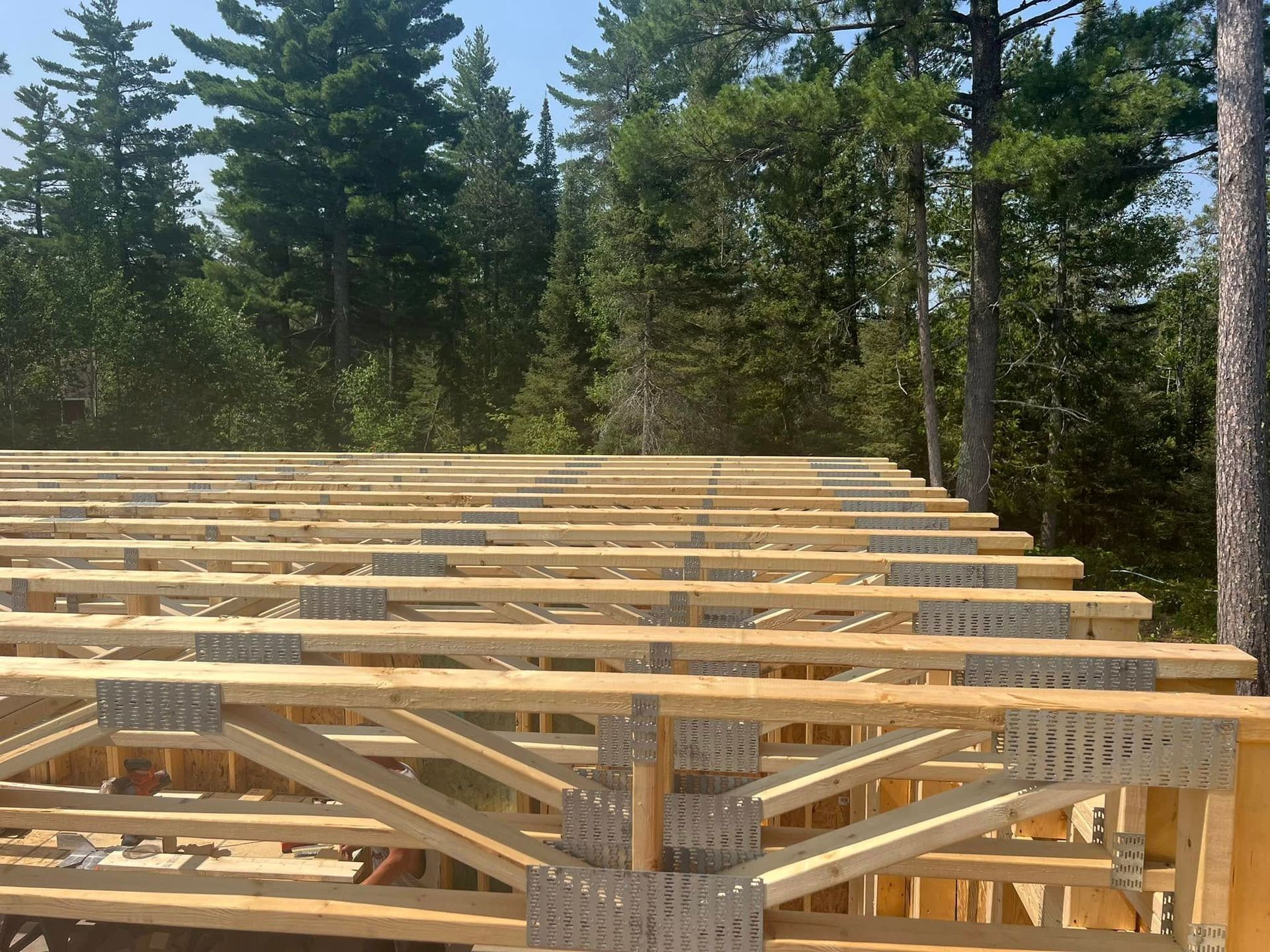 Wooden house frame under construction in a wooded area, with stacks of lumber and timber trusses.