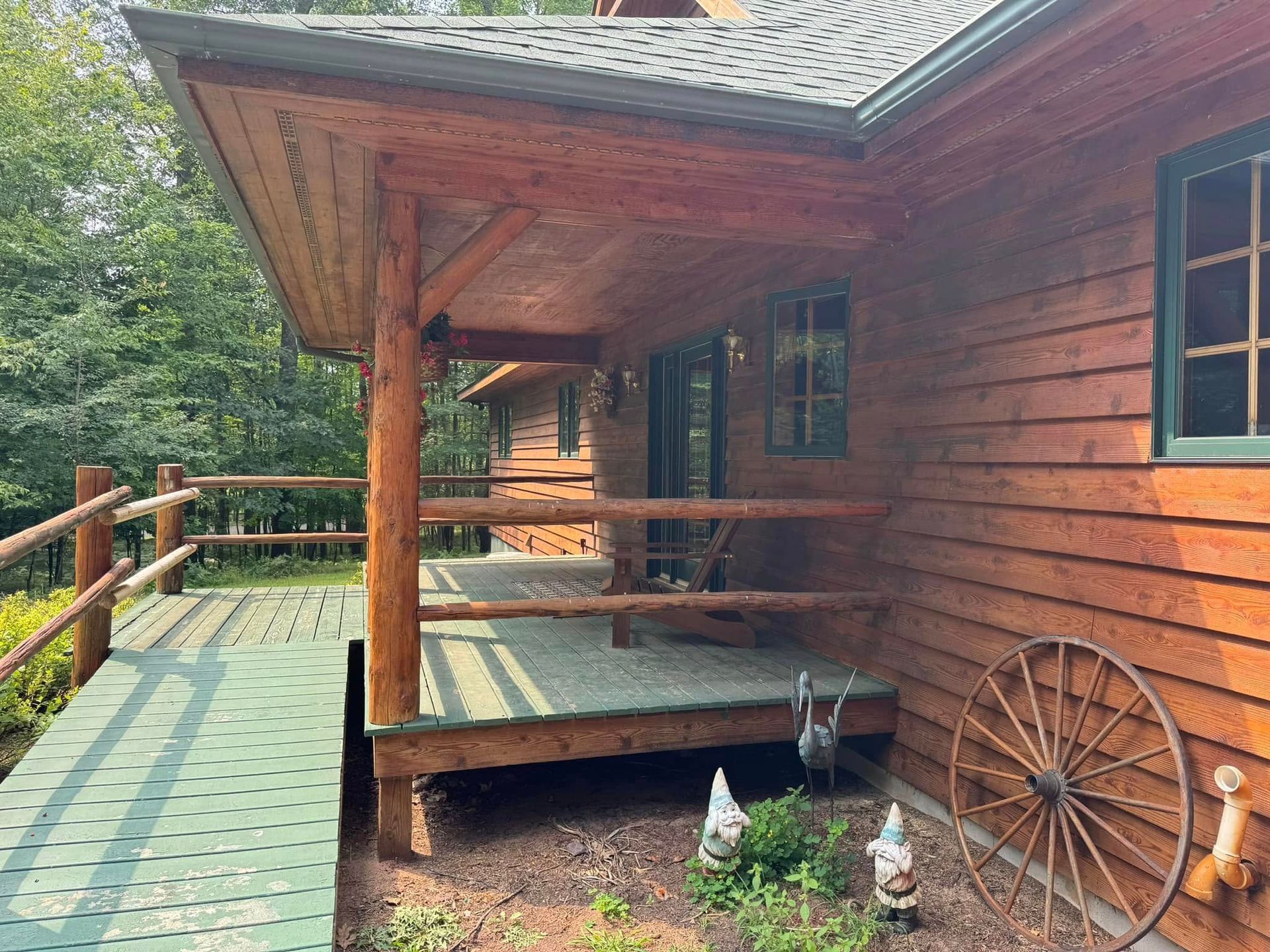 Wooden cabin porch with green roof, railing, and wagon wheel beside a forested path