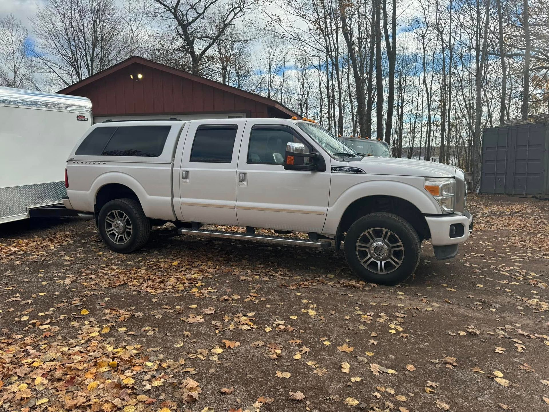 Silver pickup truck parked beside a white trailer in a leaf-covered yard near a red shed.