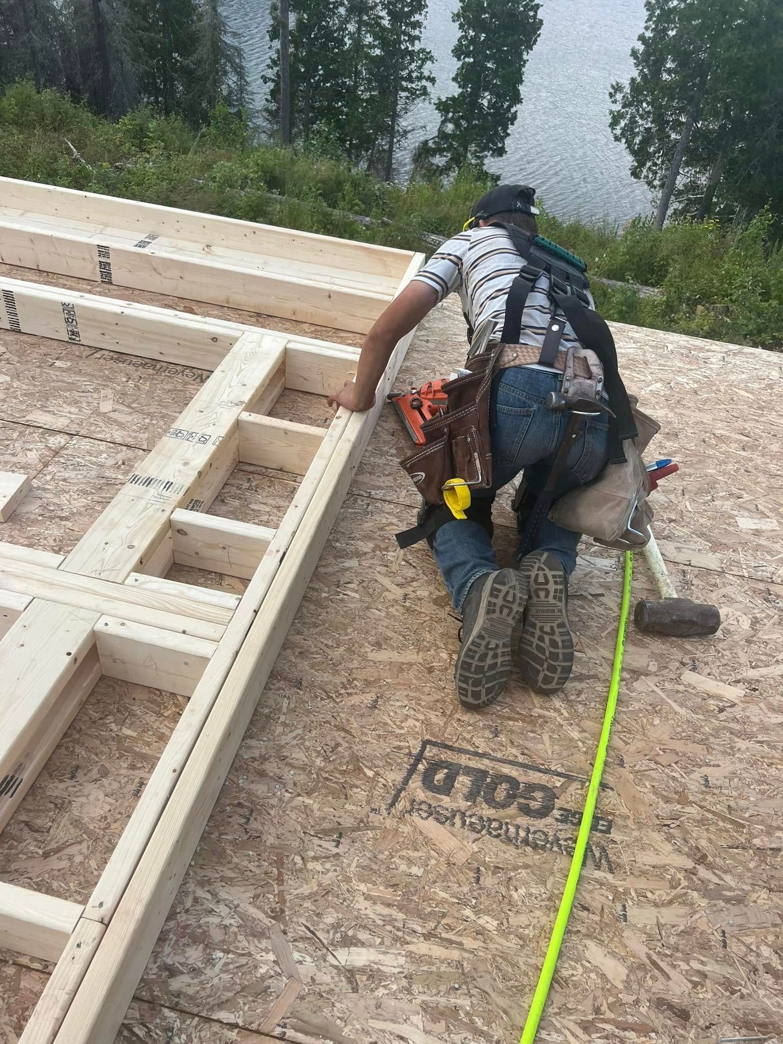 Worker kneeling on a rooftop frame, measuring and installing wood sheathing with a yellow tape line.
