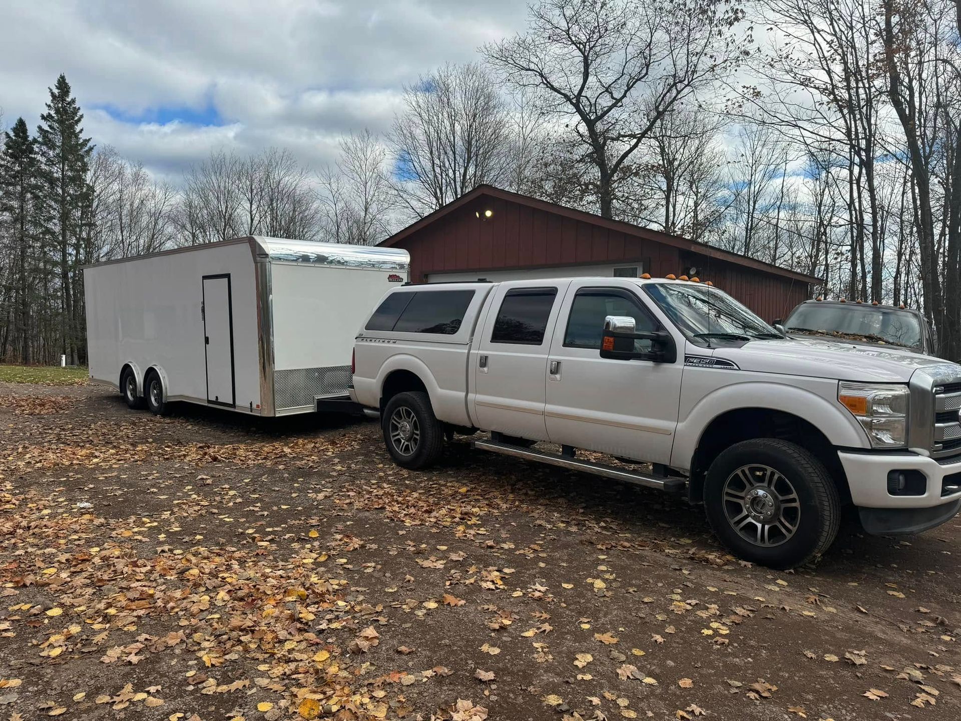 White pickup truck towing a white enclosed trailer on a leaf-covered dirt road in a wooded area
