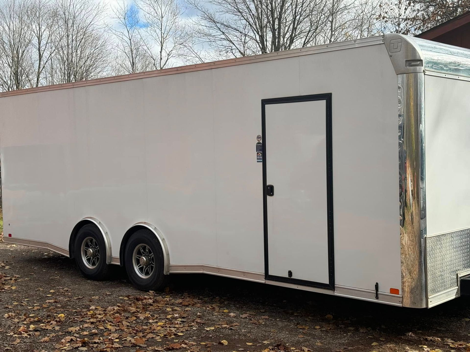 White enclosed dual-axle trailer parked outdoors on a leaf-covered lot, with a side door and silver front trim