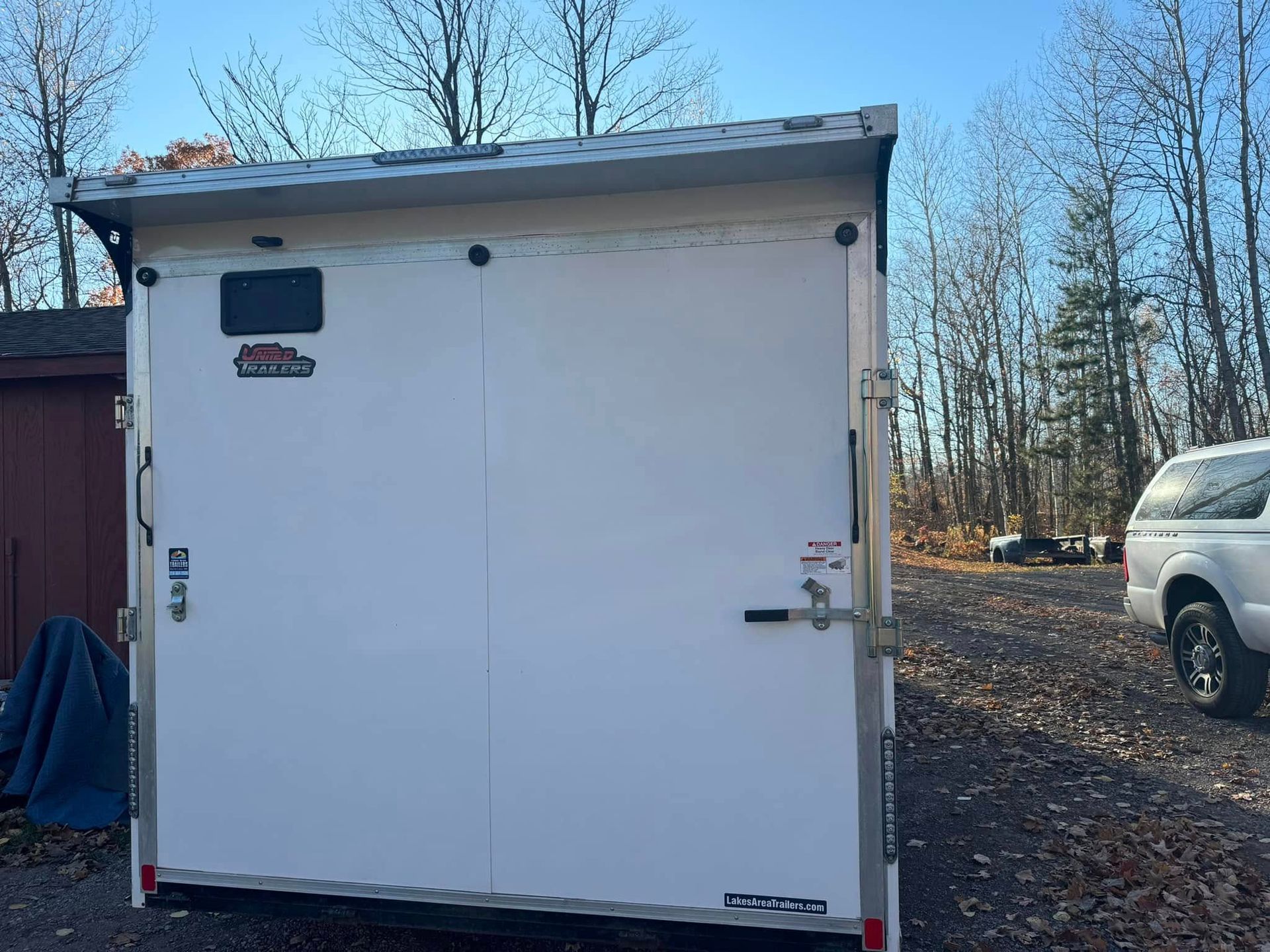 White cargo trailer parked outdoors on a gravel lot, with trees and a white SUV in the background.