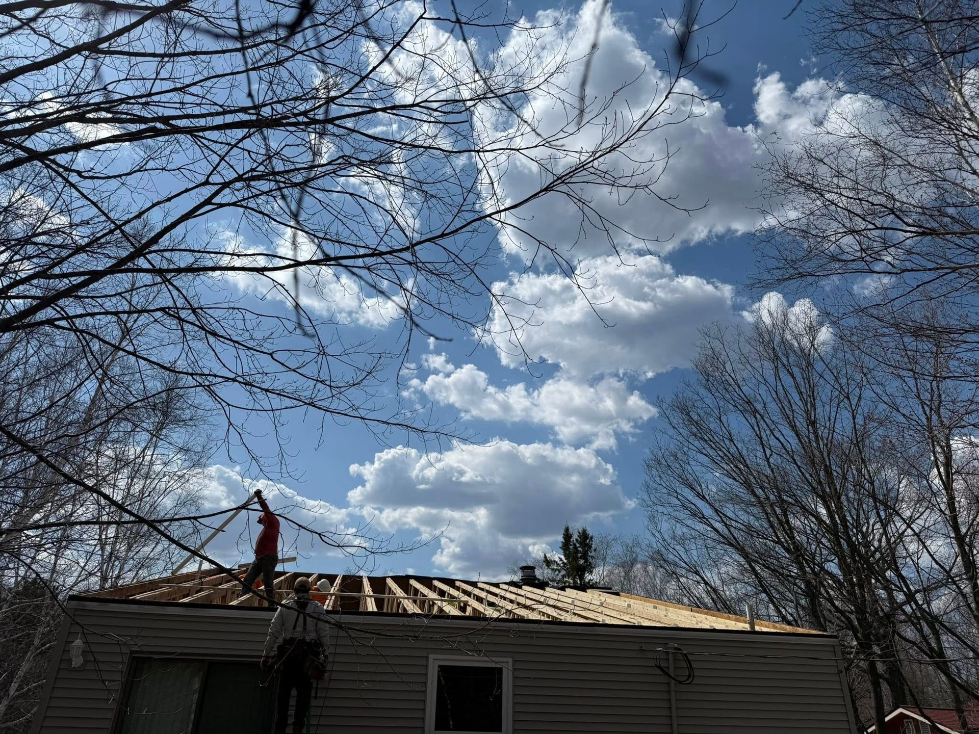 Roofing worker on a house under a cloudy blue sky with bare tree branches overhead