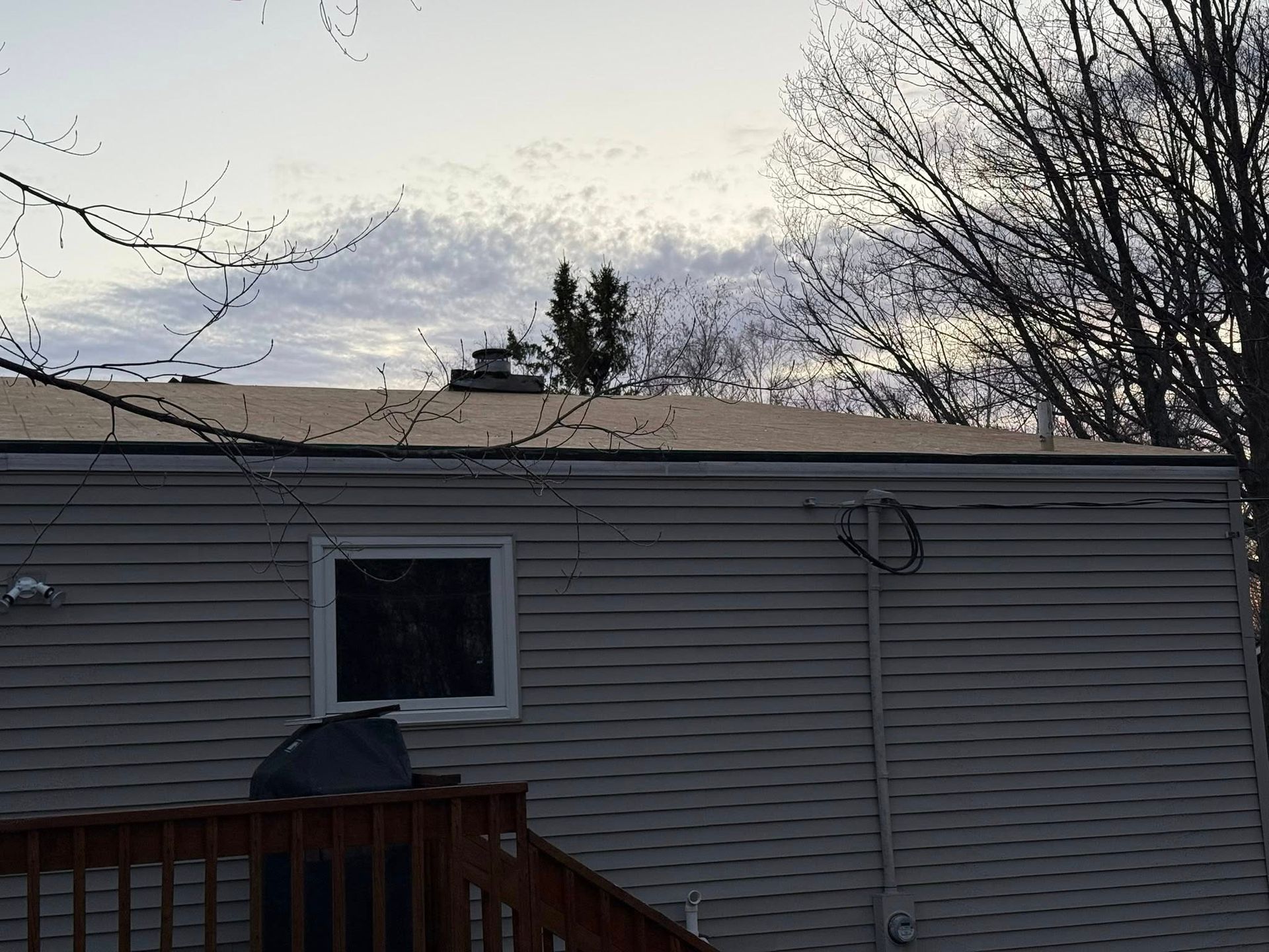 A gray-sided building with a small window, rooftop vent, and bare tree branches against a cloudy sky.