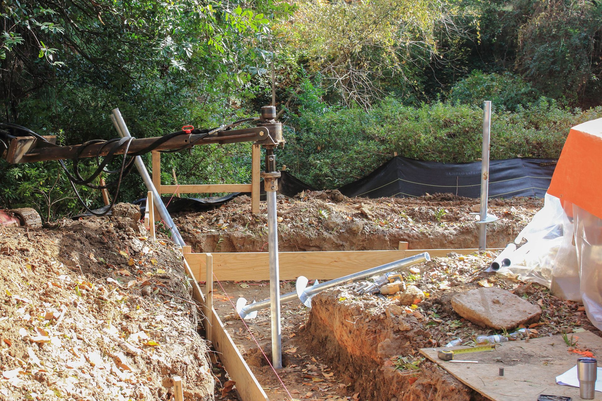 Outdoor construction site with wooden formwork, dirt piles, and a muddy trench near vegetation
