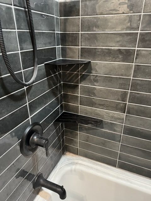 Bathroom shower corner with dark gray tile, wall-mounted fixtures, shelf, and white tub edge
