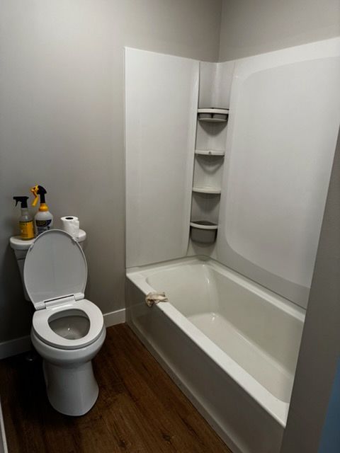 Small bathroom with toilet, bathtub, shower shelves, and cleaning supplies on the tank.