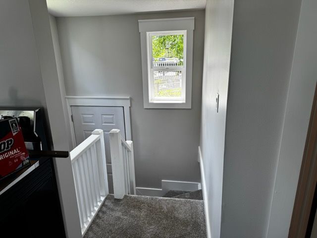 Stair landing in a gray hallway with a white railing, window, and small door opening.