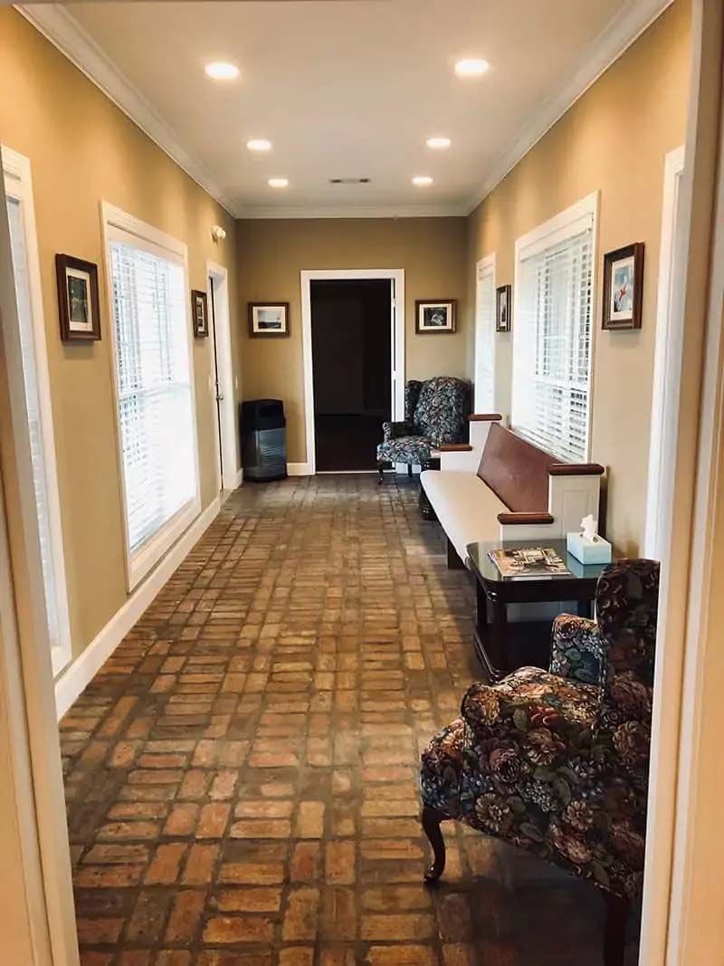 Long hallway with brick flooring, windows, and seating. Beige walls, recessed lighting.
