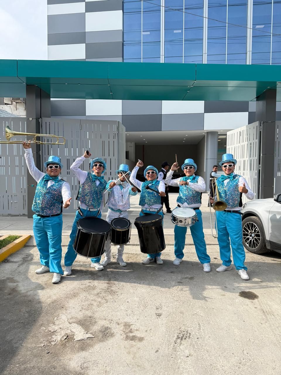 Un grupo de seis músicos, vestidos con trajes y sombreros azules, se encuentran frente a un edificio tocando instrumentos