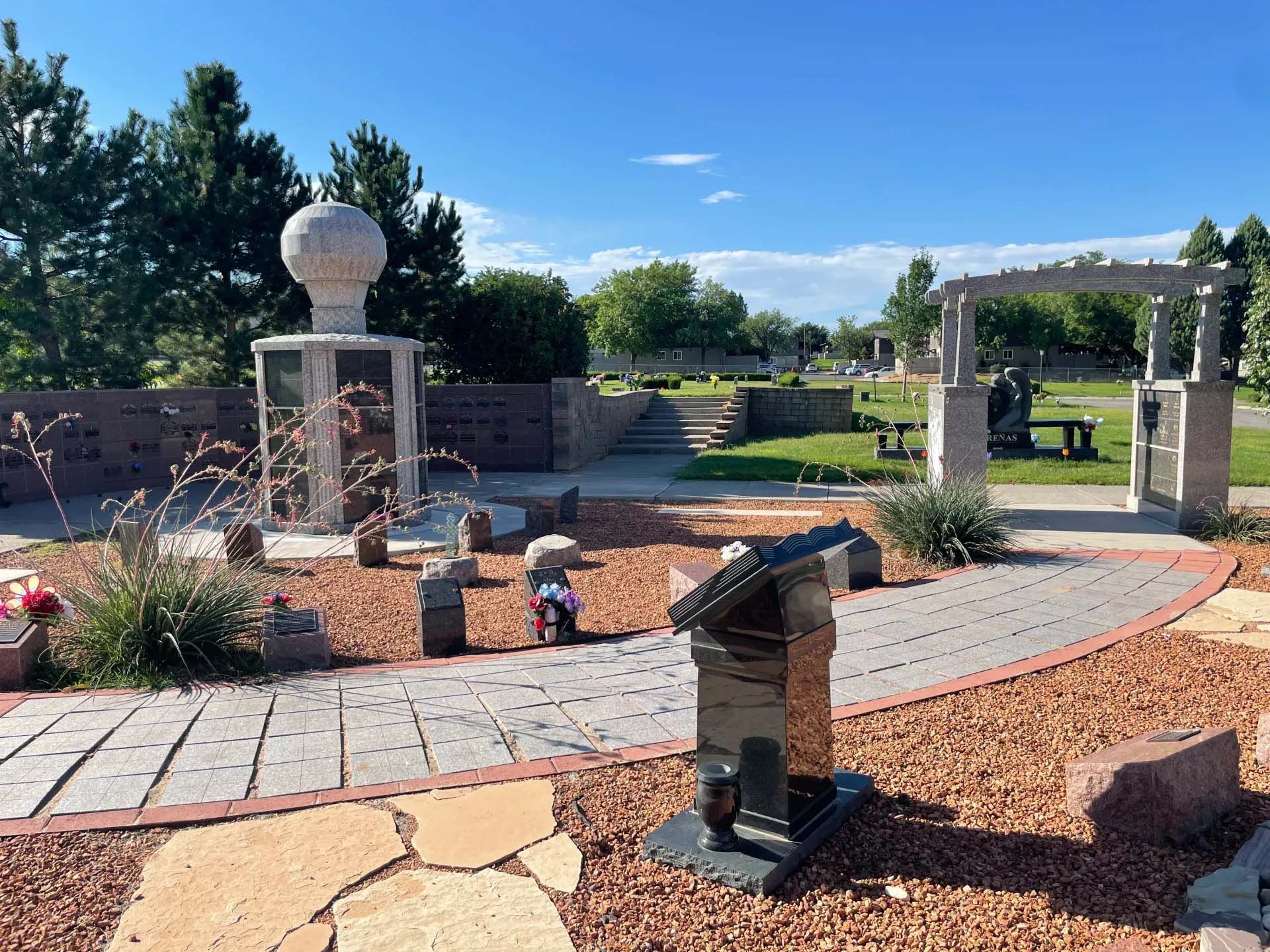 Cemetery scene with headstones, walkways, trees, and blue sky.