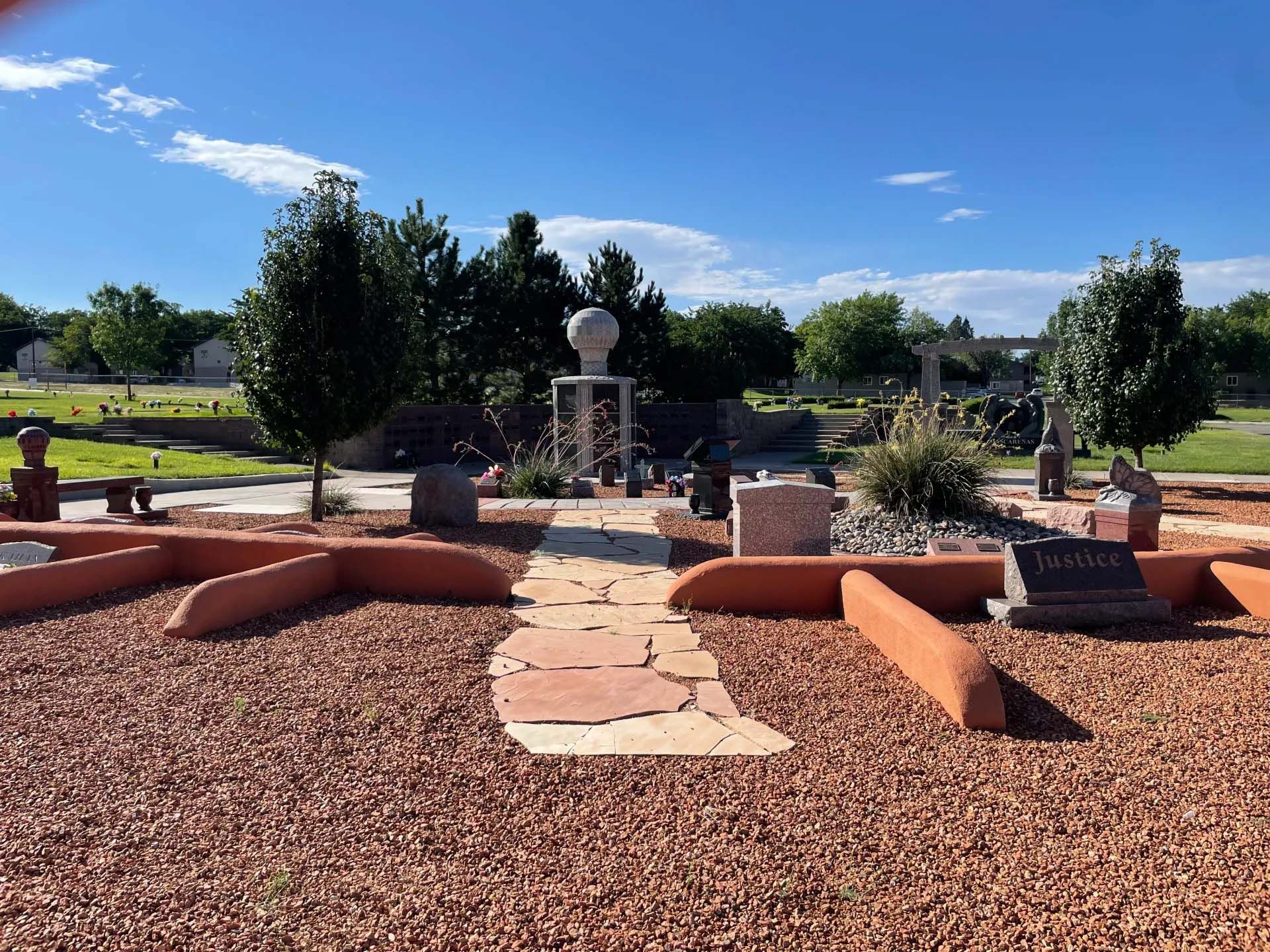 Gravel-covered cemetery with a stone pathway leading to monuments and trees under a blue sky.