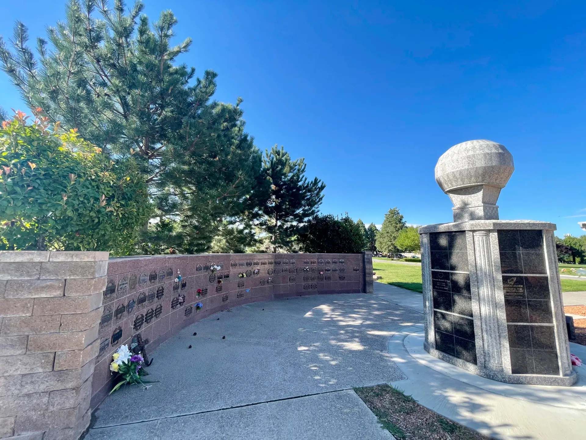Curving brick wall with engraved names, leading to a memorial column with urn-like top under a blue sky.