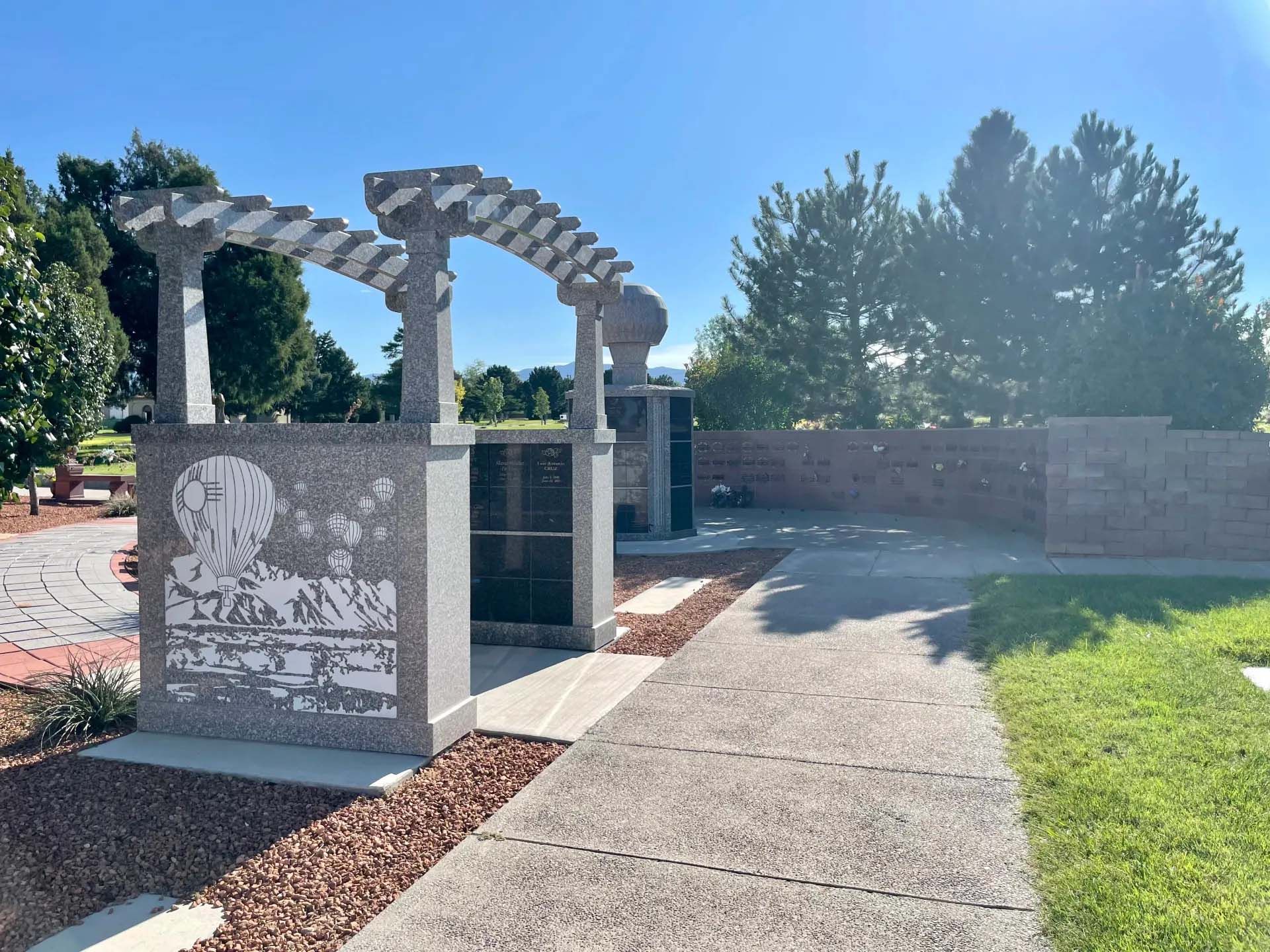 Memorial with granite structures, a pergola, and engraved panels under a clear blue sky.