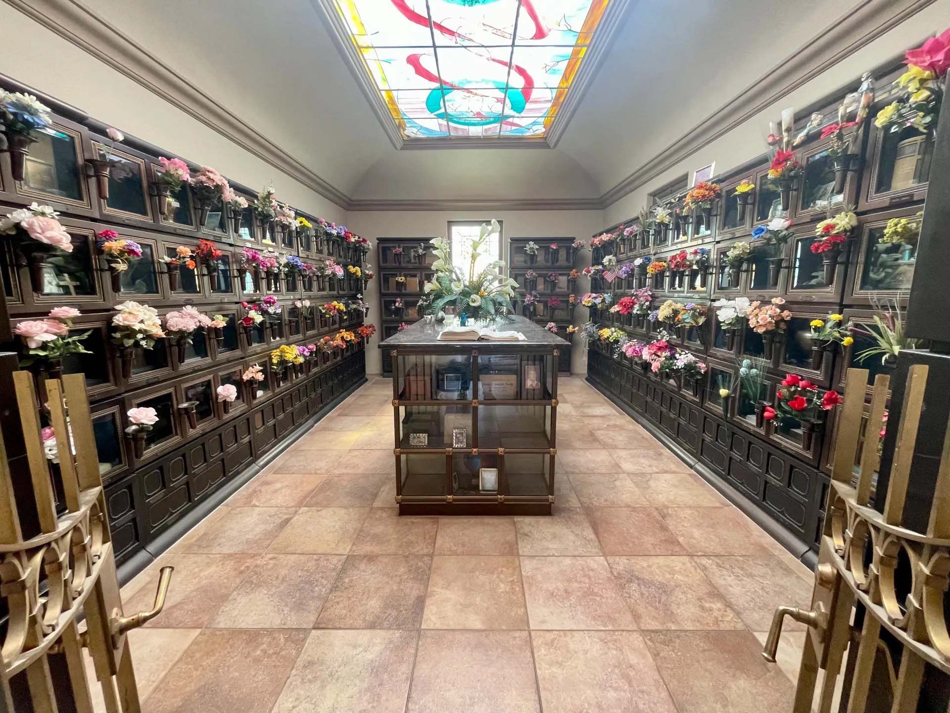 Indoor mausoleum with rows of crypts filled with flowers; stained-glass ceiling and central display.