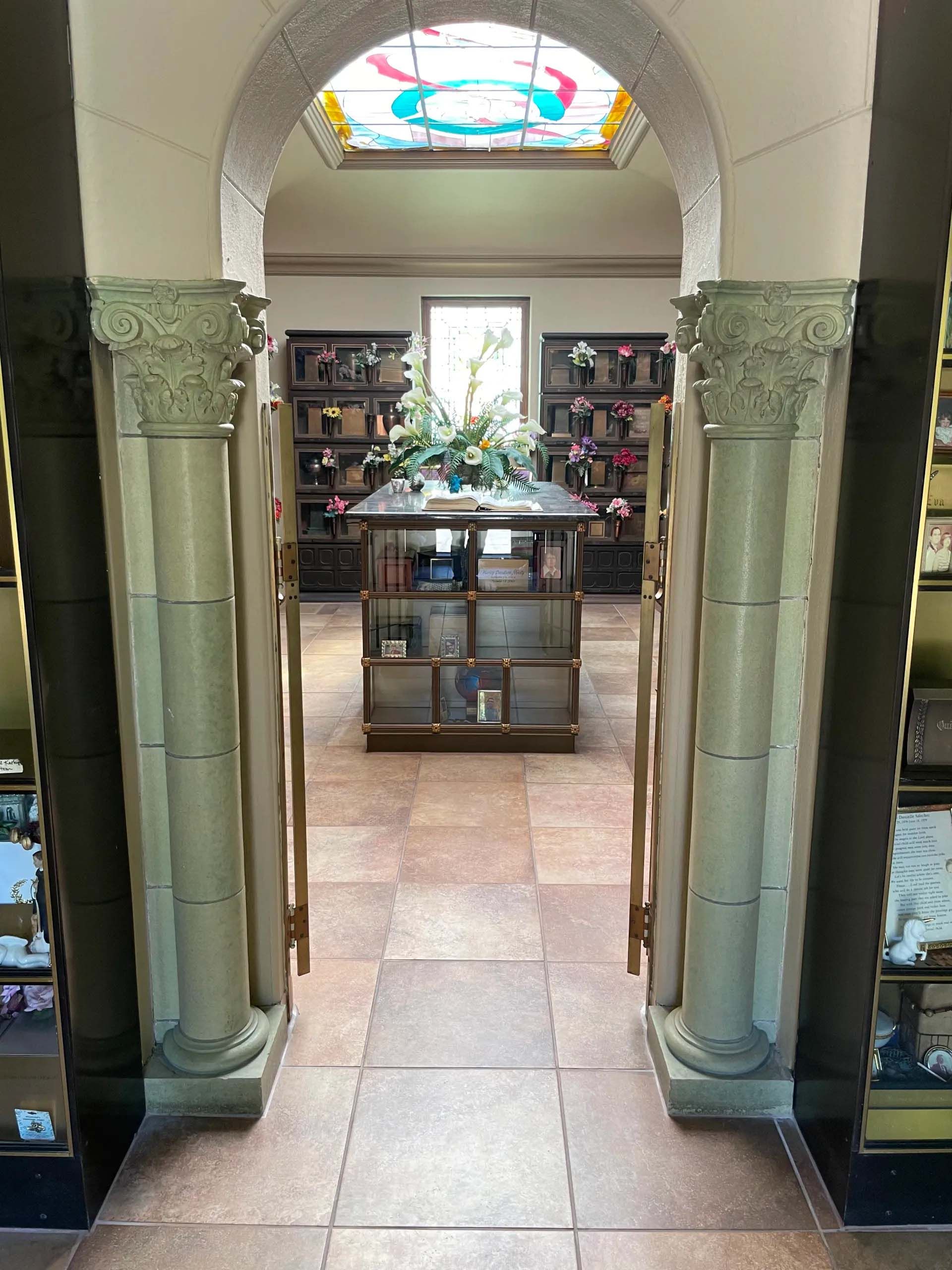 Interior of a mausoleum. Arched doorway frames a room with columns, displays, and niches for urns and flowers.