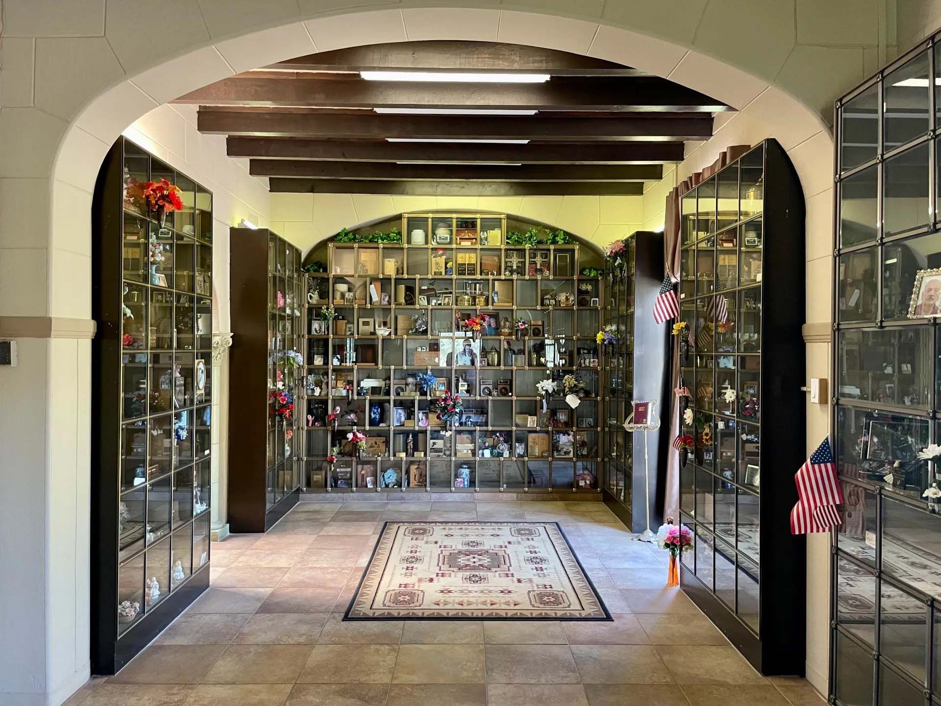 Interior of a columbarium with niches containing urns and mementos. American flags and a rug are present.