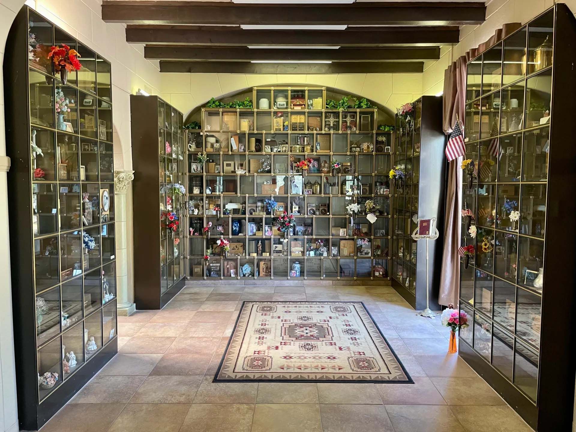 Interior of a columbarium with glass-fronted niches holding urns and memorial items. Beige stone floor, Persian rug.