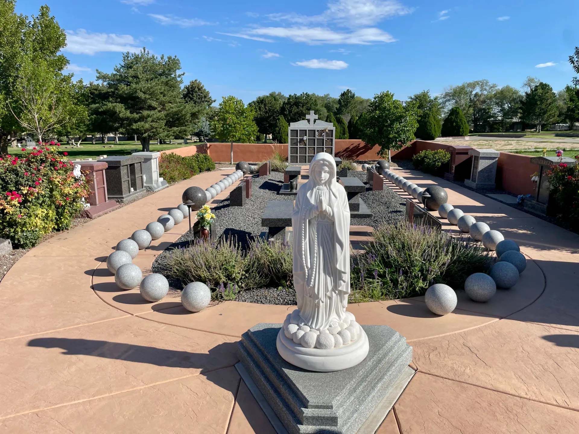 Statue of Virgin Mary in a cemetery, with rosary bead markers, greenery, and blue sky.