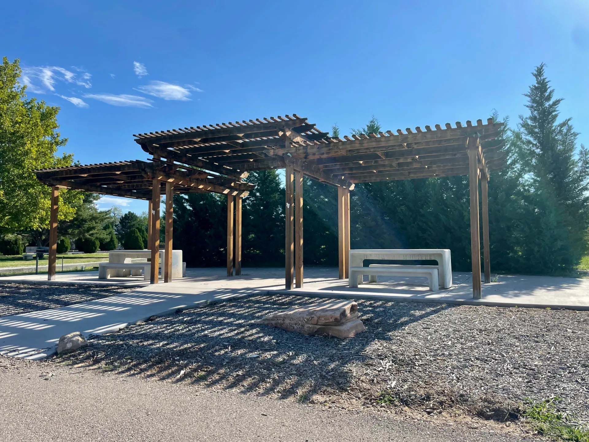 Wooden pergola with concrete benches on a gravel and paved pathway in a park.