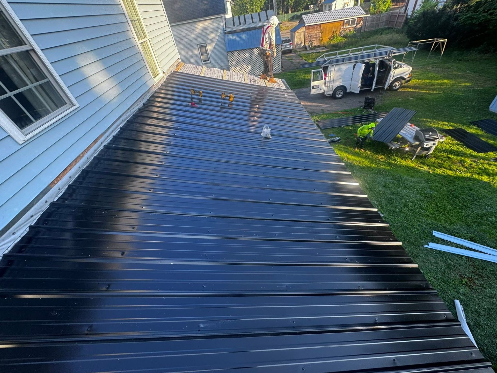 Black metal roof beside white house, viewed from above, with a person and truck in the yard below.