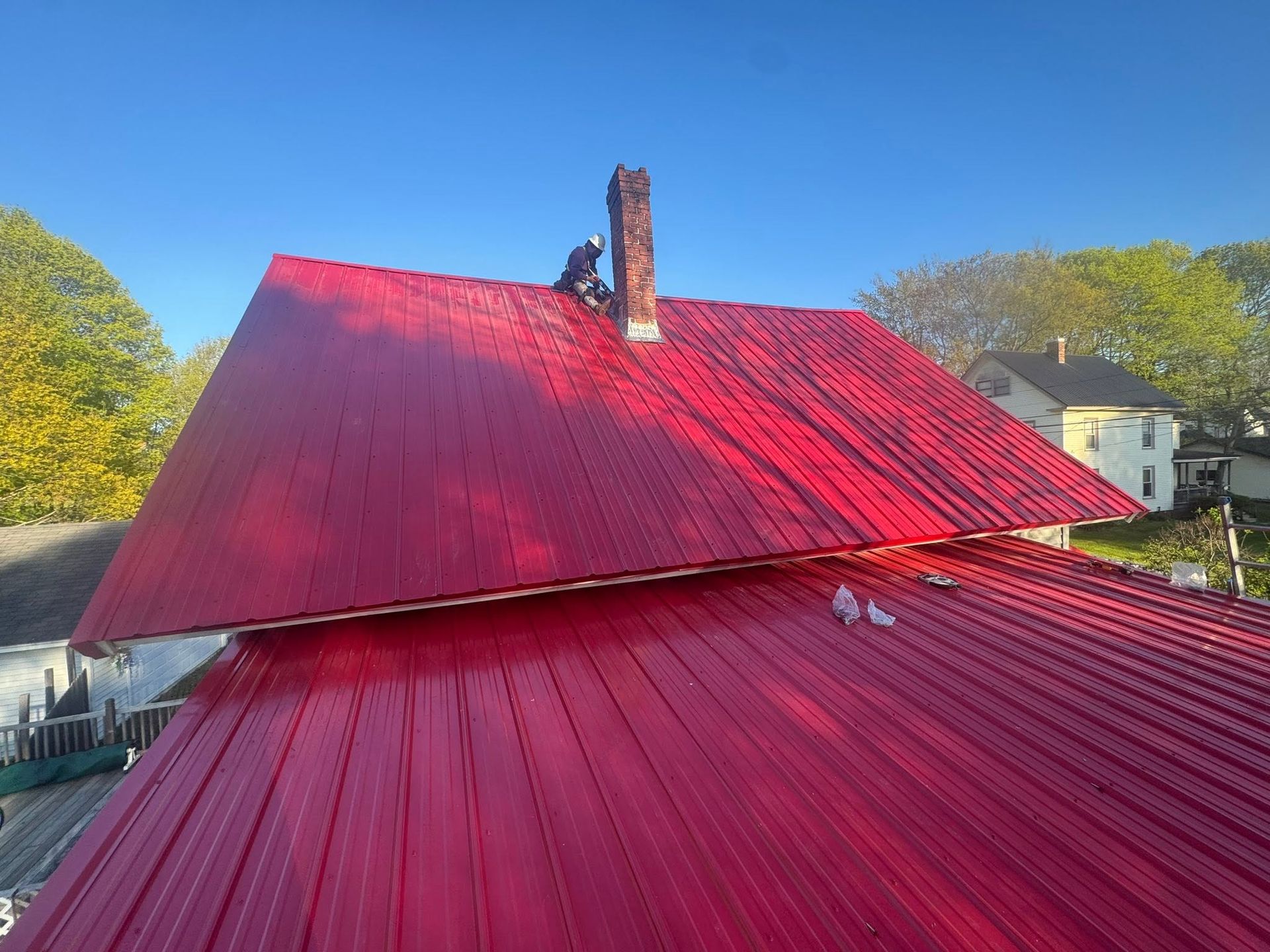Red metal roof on a house with a chimney, viewed from above against a blue sky and trees