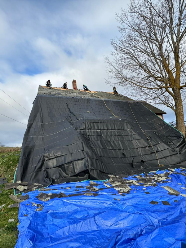 Partly covered roof on a house with black tarps and blue tarp debris in a rural yard