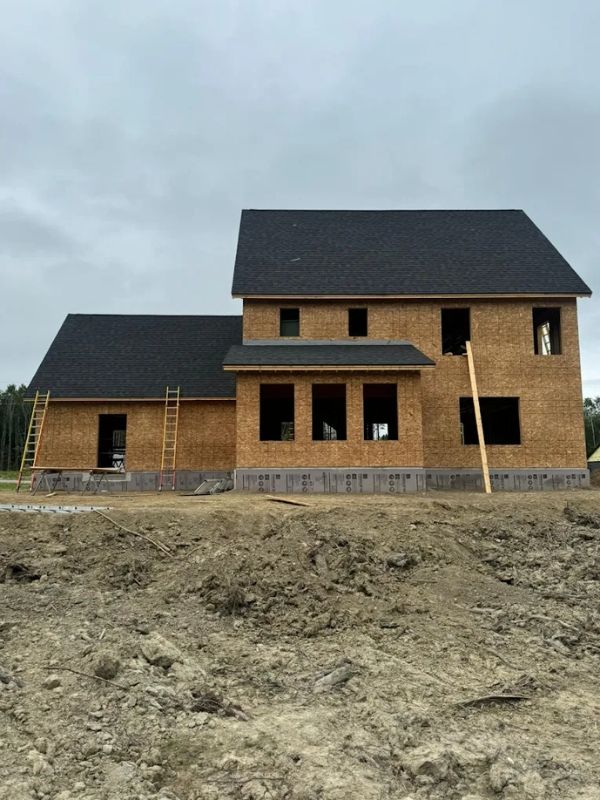 Partially built brick house with dark roof on a bare dirt lot under an overcast sky