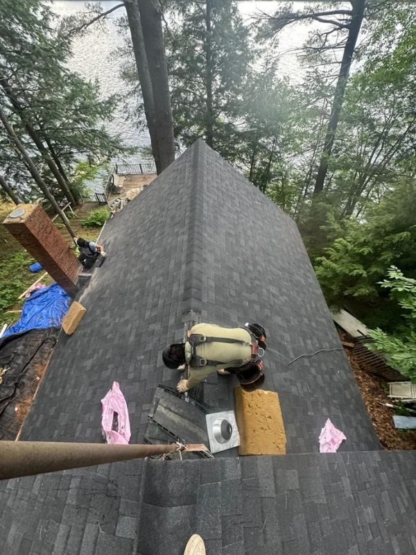 Worker installing dark gray shingles on a steep roof surrounded by trees