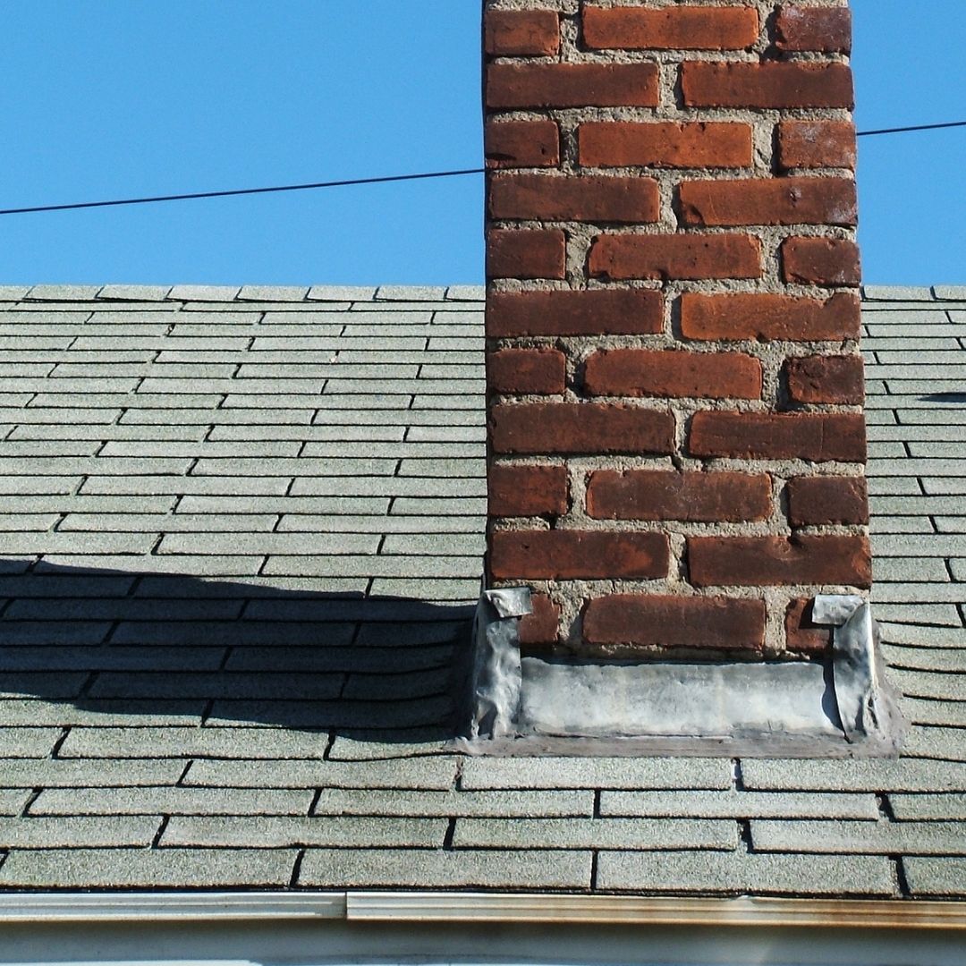 Brick chimney on a gray shingled roof under a blue sky, with a dark shadow across the roof.