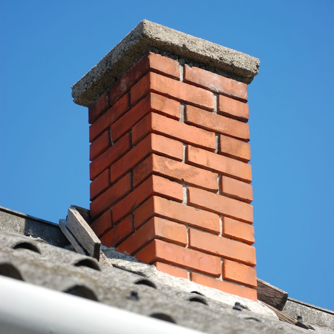 Red brick chimney with a stone cap on a gray tiled roof against a blue sky