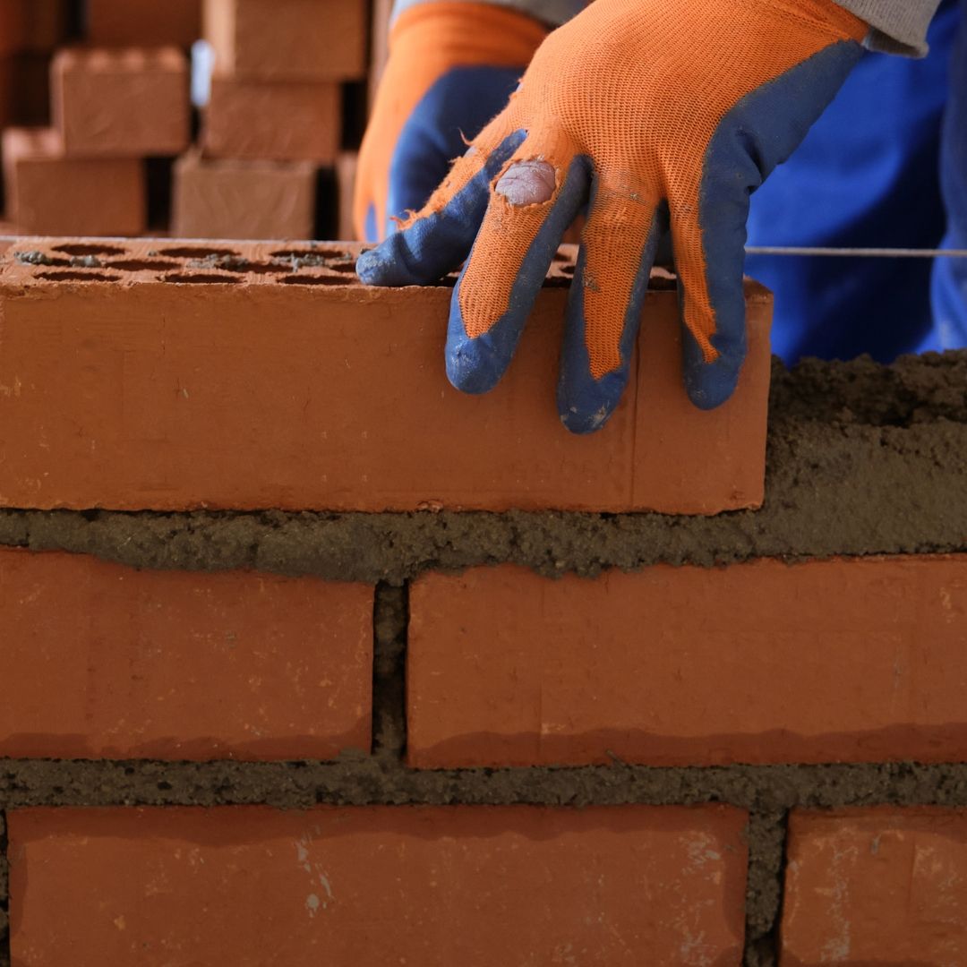 Gloved hand placing a red brick on a brick wall with wet mortar