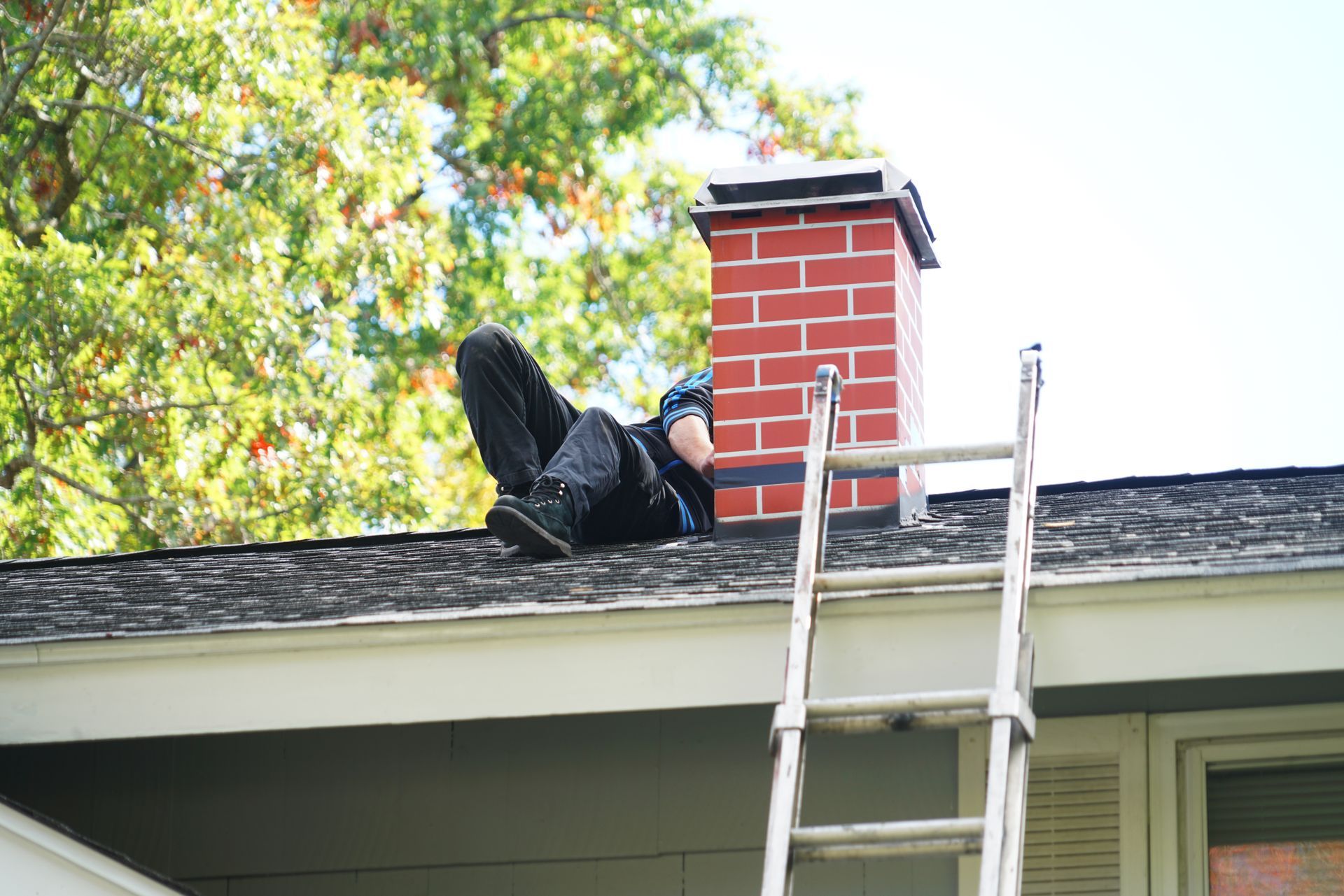 Person on a roof near a brick chimney and ladder, working on the shingles.