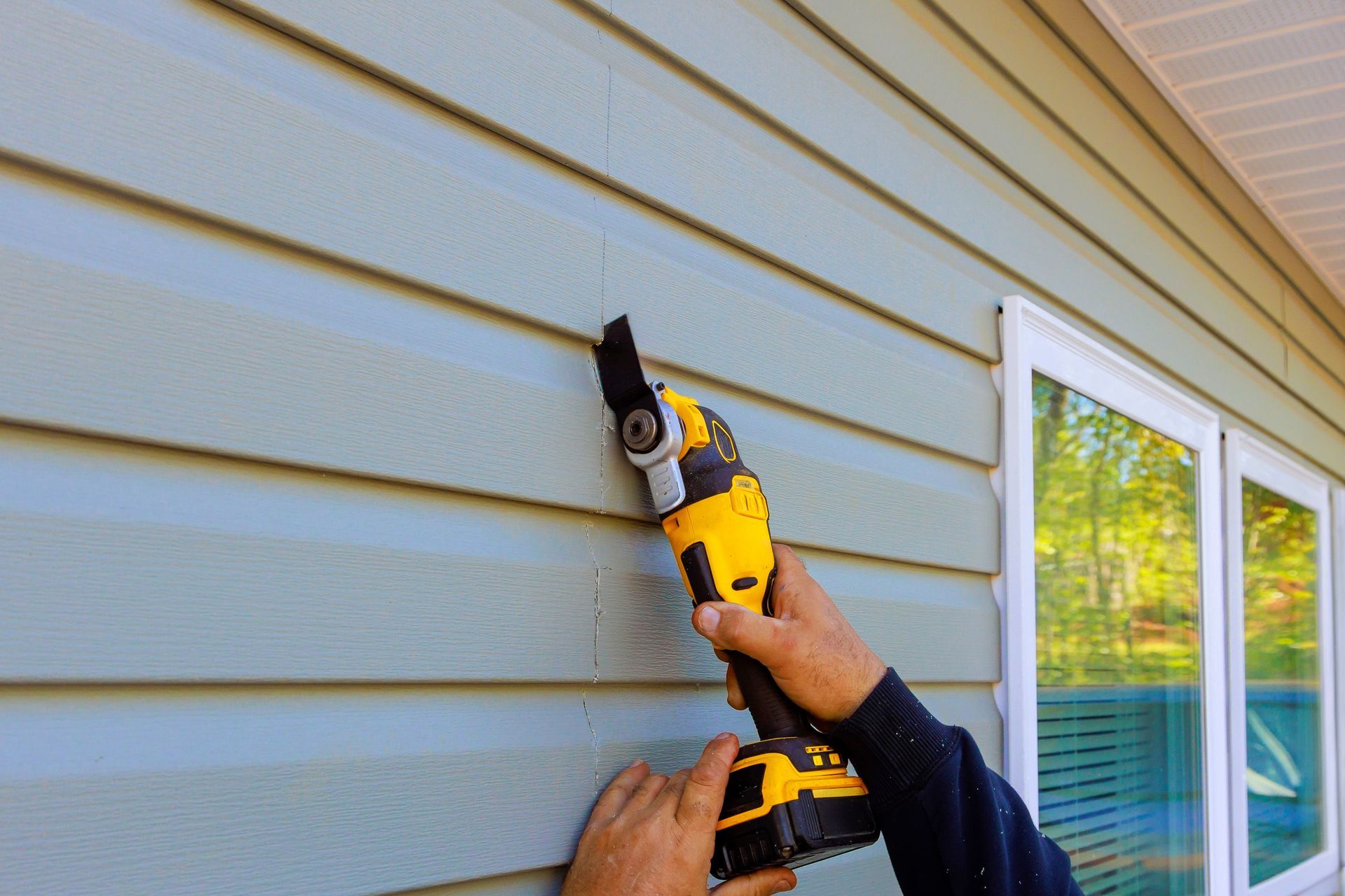 Hands using a yellow power tool to cut siding on a house exterior