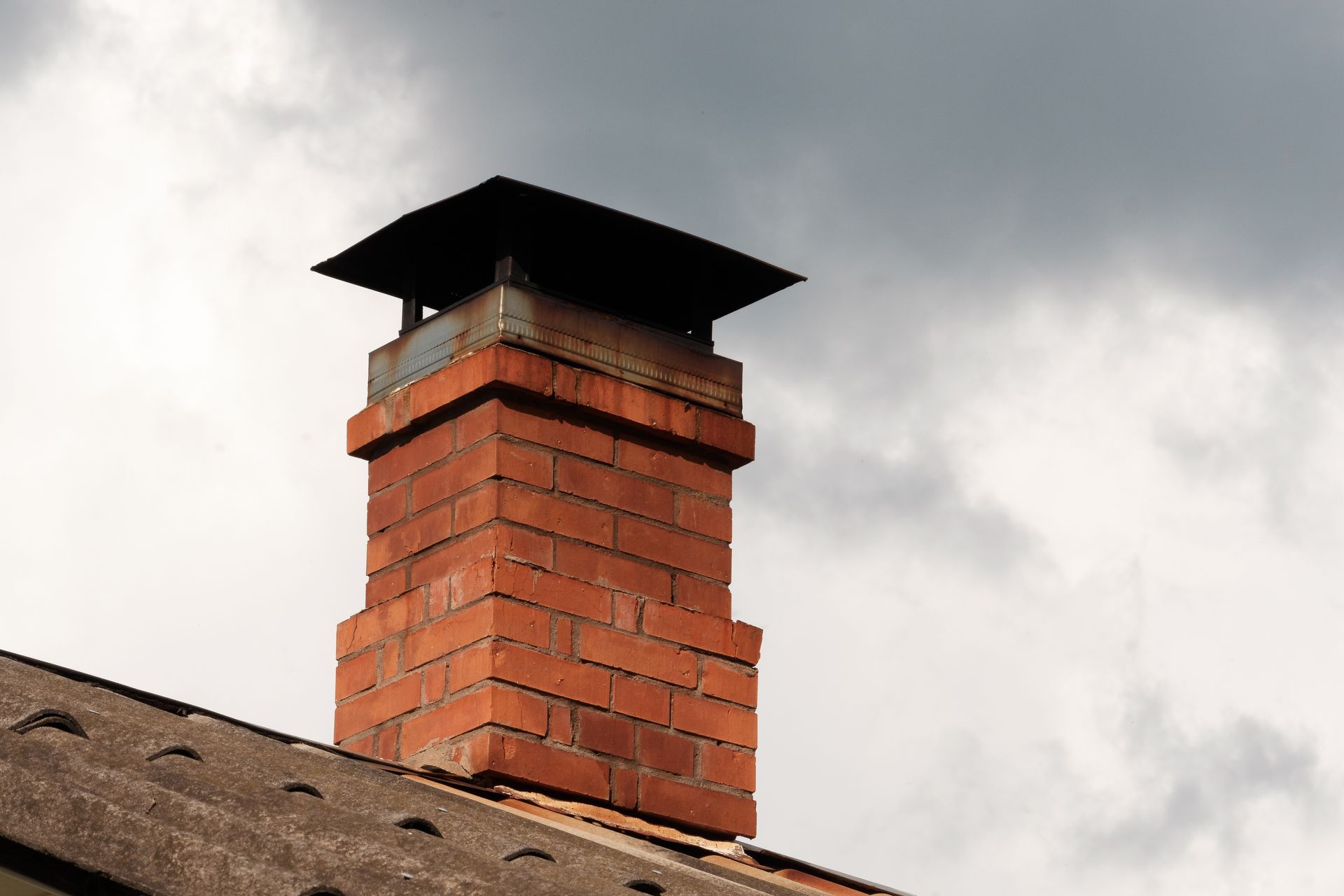 Red brick chimney with black cap on a shingled roof against a cloudy sky