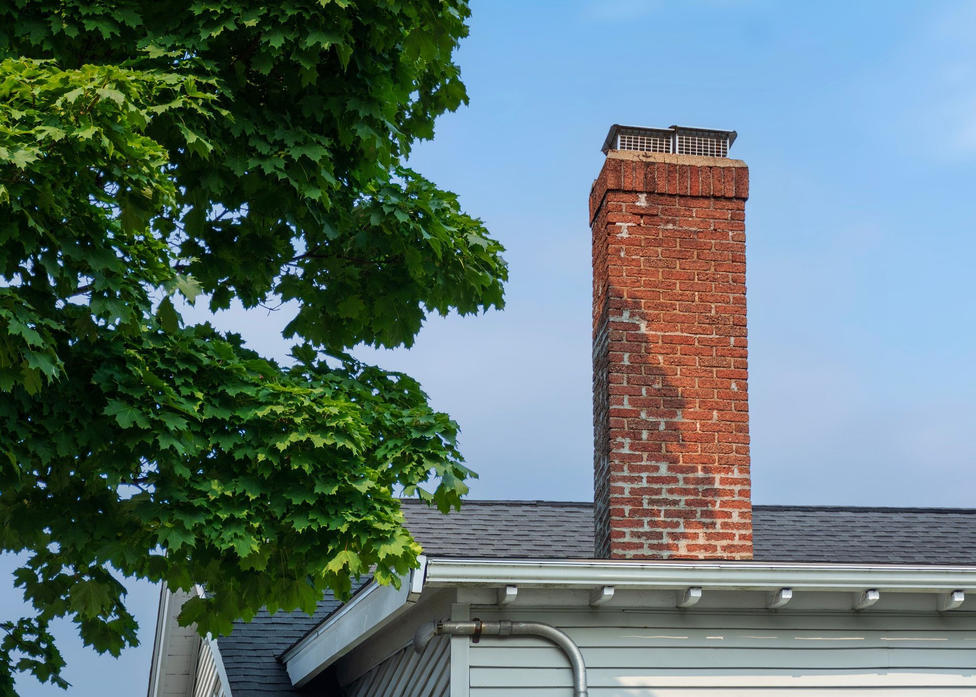 Brick chimney above a white-roofed house, partly framed by green tree leaves against a blue sky.