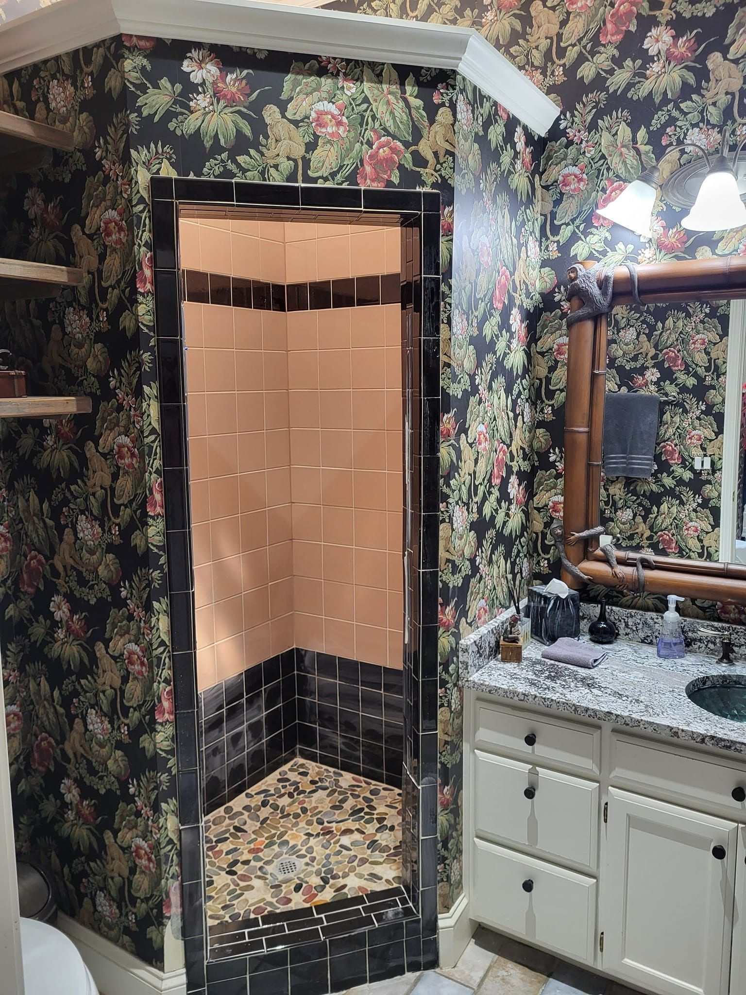Bathroom with floral wallpaper, black-tiled shower, white vanity, and stone floor.