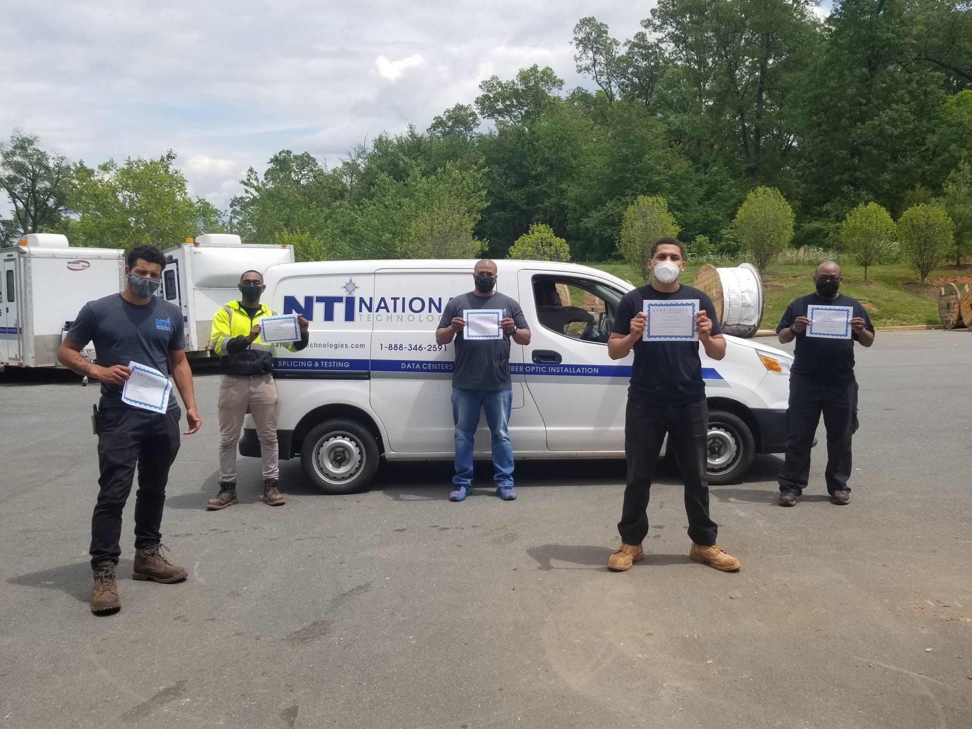 Several NTI technicians stand in front of NTI vehicles holding certificates of completion after finishing NTI's cable placement bootcamp