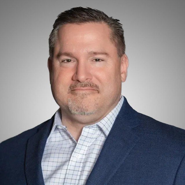 A man with a beard is wearing a blue suit and a plaid shirt.