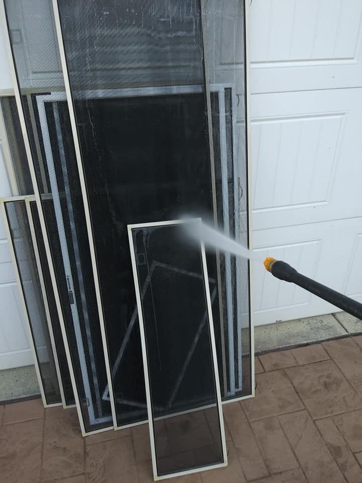 Person spraying a stack of window screens with water from a pressure washer outdoors.
