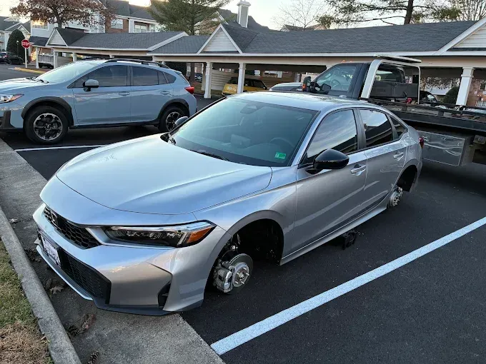 Silver sedan with a missing front wheel parked in a lot, with another car and covered parking nearby