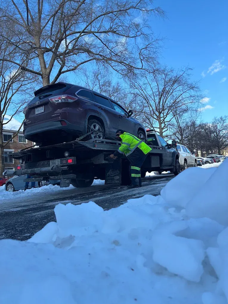 Tow truck lifting a dark SUV in snowy street while worker in neon jacket guides it