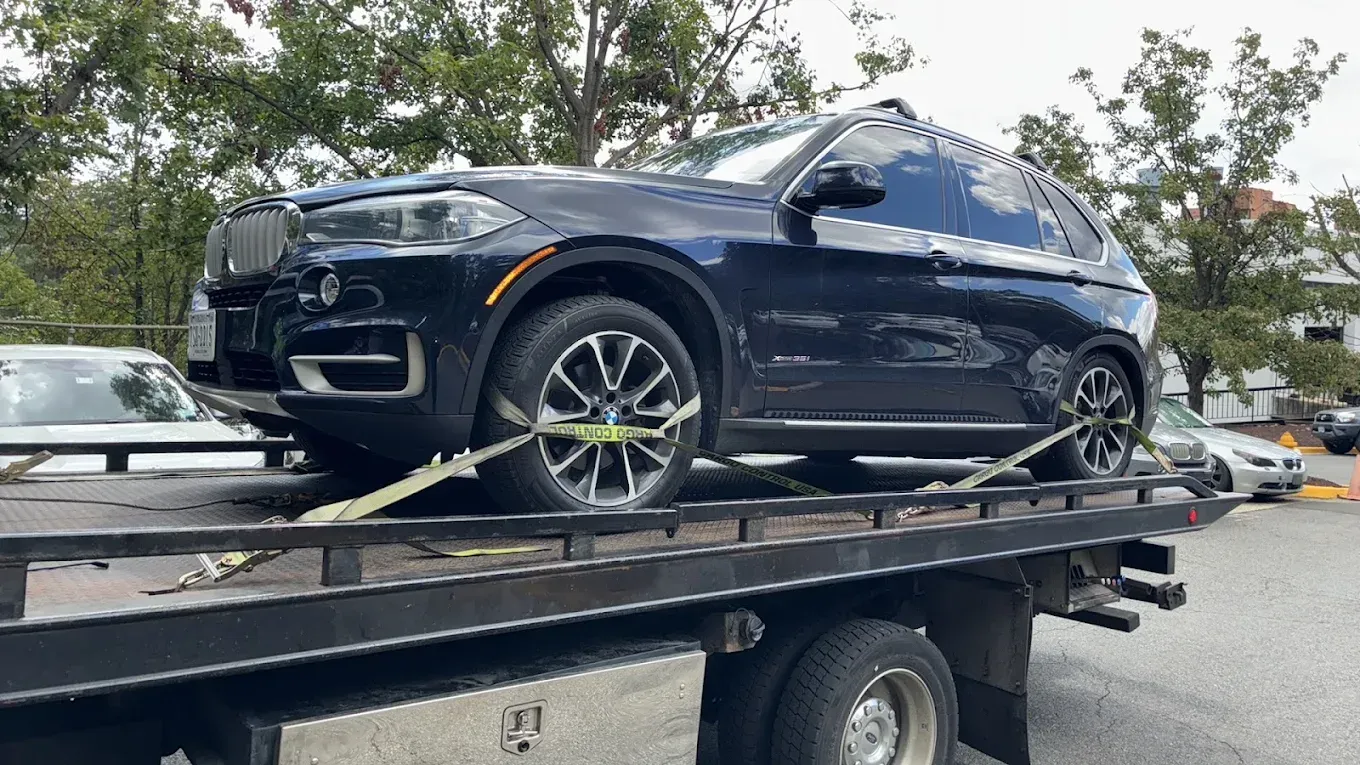 Dark blue SUV strapped to a tow truck in a parking lot, with trees and parked cars in the background.